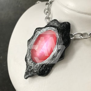 Pode incluir: Um colar de corrente prateada com um pingente preto que apresenta uma grande gema rosa iridescente. O pingente tem um design detalhado e texturizado.