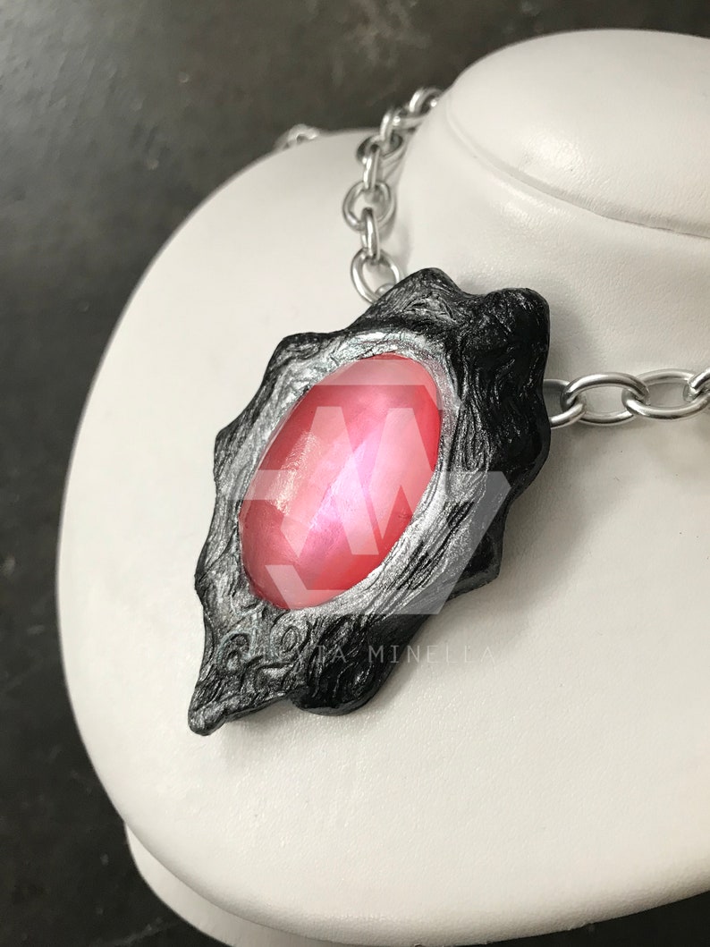 Pode incluir: Um colar de corrente prateada com um pingente preto que apresenta uma grande gema rosa iridescente. O pingente tem um design detalhado e texturizado.