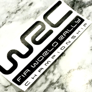 May include: A white sticker featuring the black WRC logo and the text "FIA WORLD RALLY CHAMPIONSHIP". The sticker has a rectangular shape with a black border and is set against a marble background.