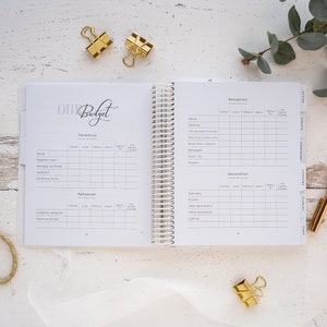 May include: A white wedding budget planner with a spiral binding. The planner has sections for ceremony, rehearsal, reception, and decoration. Each section has columns for estimated cost, actual cost, difference, deposit, balance, and final due date.