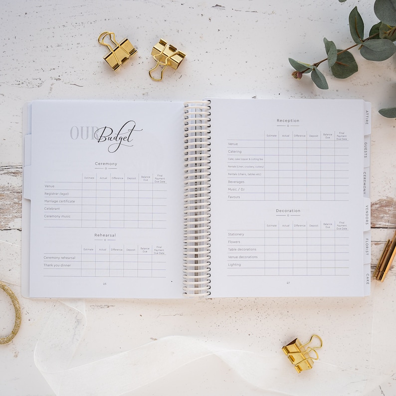 May include: A white wedding budget planner with a spiral binding. The planner has sections for ceremony, rehearsal, reception, and decoration. Each section has columns for estimated cost, actual cost, difference, deposit, balance, and final due date.