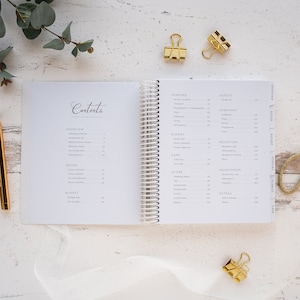 May include: A white wedding planner with a spiral binding. The planner has tabs for different sections, including Vendors, Guests, Ceremony, Reception, Honeymoon, Attire, Florist, Cake, Decor, Budget, and Extras.