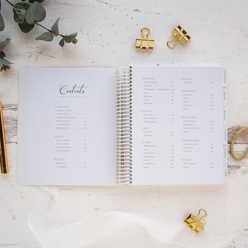 May include: A white wedding planner with a spiral binding. The planner has tabs for different sections, including Vendors, Guests, Ceremony, Reception, Honeymoon, Attire, Florist, Cake, Decor, Budget, and Extras.