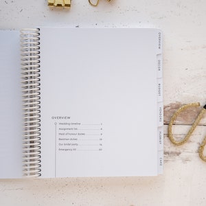 May include: A white wedding planner with a spiral binding and tabs for different categories, including overview, decor, budget, vendors, florist, and cake. The planner has a list of tasks with corresponding page numbers.