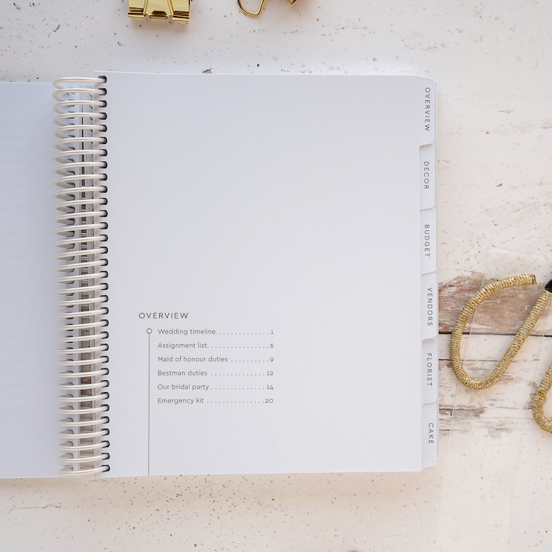 May include: A white wedding planner with a spiral binding and tabs for different categories, including overview, decor, budget, vendors, florist, and cake. The planner has a list of tasks with corresponding page numbers.