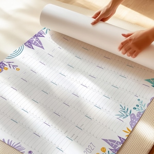 Puede incluir: Un calendario de pared grande, blanco y desplegado para el a&ntilde;o 2027. El calendario presenta un dise&ntilde;o de cuadr&iacute;cula con columnas para los meses y filas para los d&iacute;as. Est&aacute; decorado con ilustraciones estilizadas de flores y hojas en morado, amarillo y verde azulado.