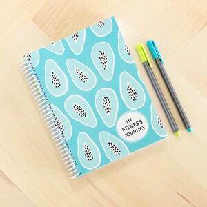 Puede incluir: Una libreta azul de espiral con un patrón de papaya y el texto "My Fitness Journey". La libreta está sobre una superficie de madera clara con dos bolígrafos, uno verde y uno negro.