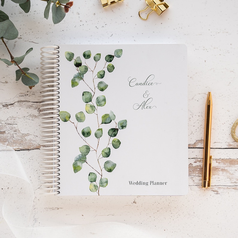 May include: A white wedding planner featuring a watercolour illustration of eucalyptus leaves. The planner has the names "Candice & Alex" and the text "Wedding Planner" printed on the cover.