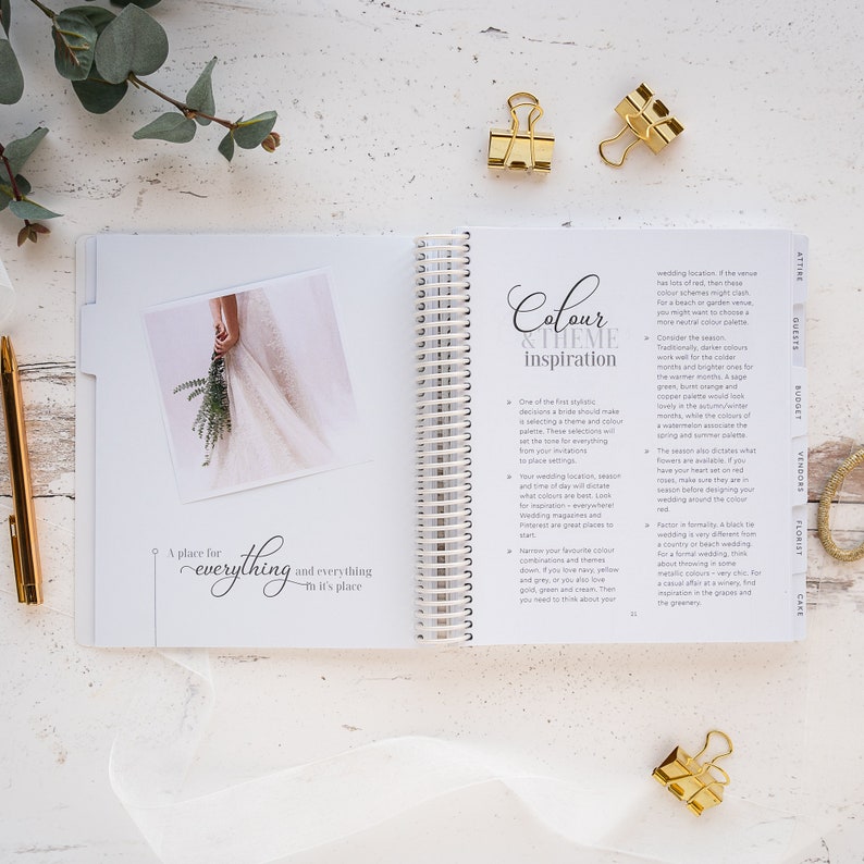 May include: An open notebook with a white background and gold spiral binding. The notebook has the words "Colour & Theme Inspiration" on the cover. The page is open to a section about choosing a wedding colour scheme. The text on the page includes tips for selecting colours based on the wedding location, season, and formality.