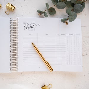 May include: An open spiral bound notebook with a white cover and the words "Our Guest List" written in black cursive. The notebook has columns for name, address, number of people, relationship, invited to ceremony, accepted ceremony, invited to reception, and accepted reception. There are check boxes next to each column.