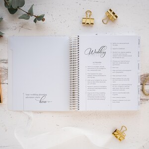 May include: A wedding planner with a white cover and gold spiral binding. The cover has the word "Wedding" written in cursive script. The planner is open to a page with a checklist of tasks to complete for planning a wedding. The checklist includes tasks such as setting a date, booking a venue, and choosing a photographer.