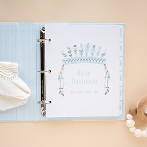 May include: A blue and white baby memory book with a decorative feather headdress and the name "Oscar Davidson" on the cover. The book has tabs for "Pregnancy", "Birth", "Firsts", "Family", "Months", and "Years".