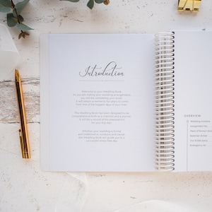 May include: A white wedding planner with a spiral binding. The cover features the word "Introduction" written in cursive. The page is open to the introduction section, which welcomes the reader to the book and explains its purpose. The book is designed to be a comprehensive checklist and planner for wedding preparations.