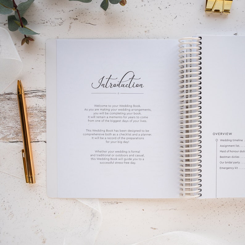 May include: A white wedding planner with a spiral binding. The cover features the word "Introduction" written in cursive. The page is open to the introduction section, which welcomes the reader to the book and explains its purpose. The book is designed to be a comprehensive checklist and planner for wedding preparations.