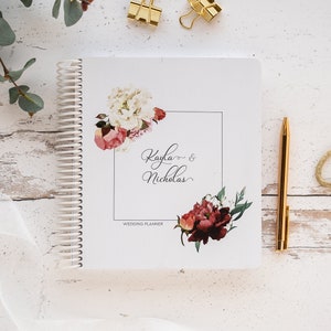 K&ouml;nnte beinhalten: Ein wei&szlig;es Hochzeitsplaner-Notizbuch mit Spiralbindung, floralen Designs und den Namen "Kayla & Nicholas". Ein goldener Stift und B&uuml;roklammern liegen daneben. Der Planer ist mit Aquarellblumen in Rosa, Rot und Wei&szlig; verziert. Der Text "Wedding Planner" ist ebenfalls sichtbar.