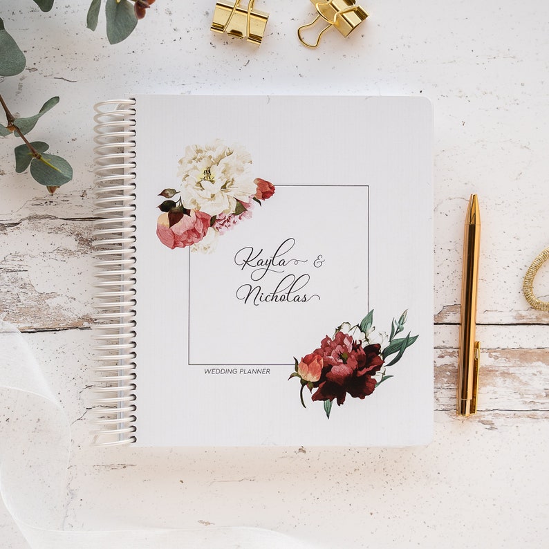 K&ouml;nnte beinhalten: Ein wei&szlig;es Hochzeitsplaner-Notizbuch mit Spiralbindung, floralen Designs und den Namen "Kayla & Nicholas". Ein goldener Stift und B&uuml;roklammern liegen daneben. Der Planer ist mit Aquarellblumen in Rosa, Rot und Wei&szlig; verziert. Der Text "Wedding Planner" ist ebenfalls sichtbar.