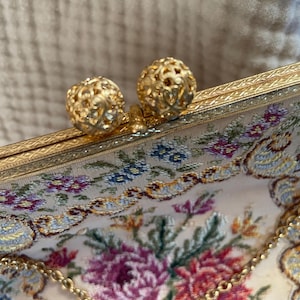 Puede incluir: Primer plano de un bolso de mano vintage de tapiz floral con herrajes dorados. El cierre presenta dos esferas caladas y ornamentadas. Una delicada cadena dorada y un estampado floral en rosa, morado, azul y verde adornan la tela.