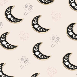 May include: A repeating pattern of crescent moon pins with a black enamel finish and gold trim. The moons feature white circles and floral details. The background is a light pink color with line drawings of hands, crystals, and eyes.
