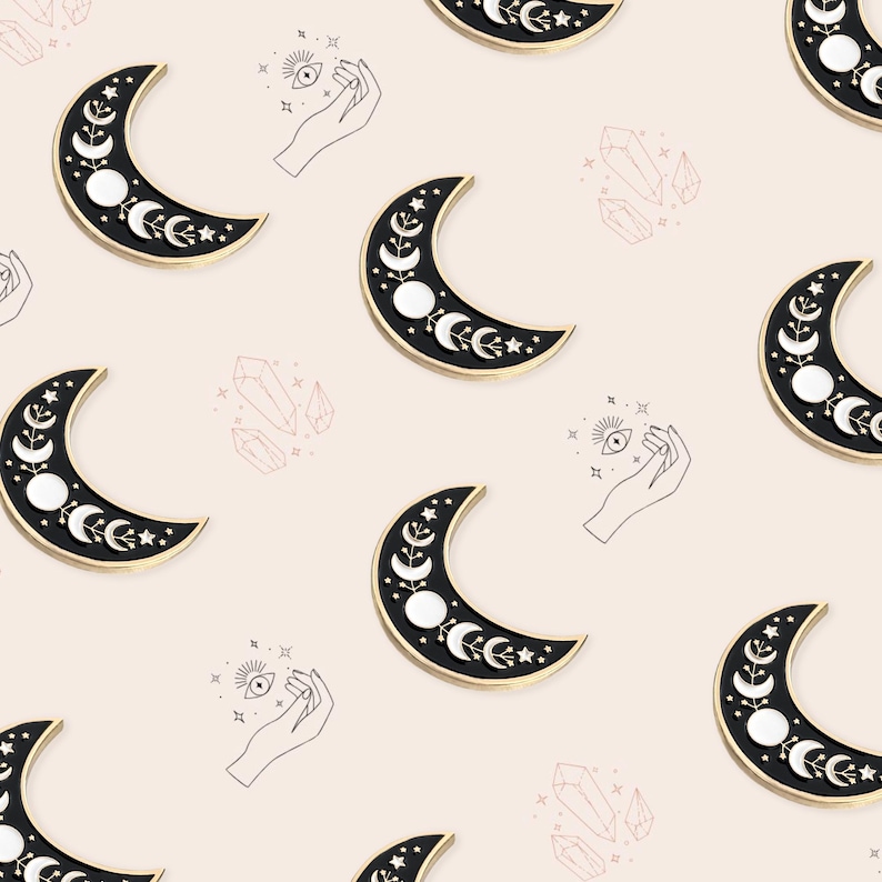May include: A repeating pattern of crescent moon pins with a black enamel finish and gold trim. The moons feature white circles and floral details. The background is a light pink color with line drawings of hands, crystals, and eyes.