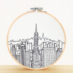 May include: A black and white embroidered cityscape design on a white fabric stretched in a wooden embroidery hoop.