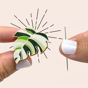 Pode incluir: Um alfinete de esmalte em forma de folha de monstera, verde, branco e dourado, com uma agulha. O design do alfinete imita uma folha com veias detalhadas. Os dedos de uma pessoa seguram o alfinete, com uma agulha no fundo.
