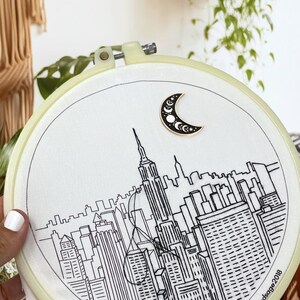 May include: Embroidery hoop featuring a black and white line drawing of a cityscape, including the Empire State Building. A crescent moon pin with stars is attached to the fabric. The hoop is cream-colored.