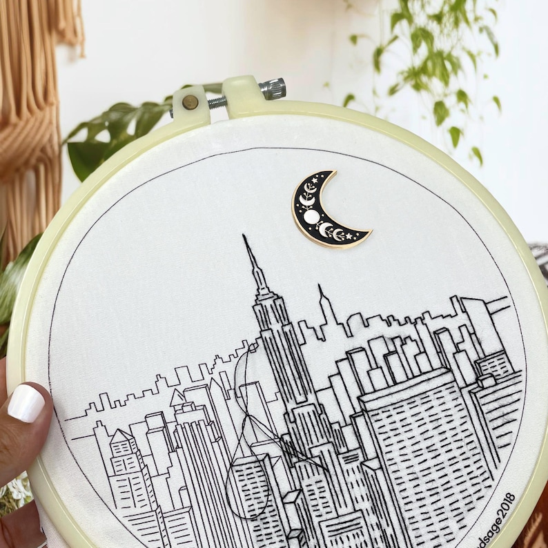 May include: Embroidery hoop featuring a black and white line drawing of a cityscape, including the Empire State Building. A crescent moon pin with stars is attached to the fabric. The hoop is cream-colored.