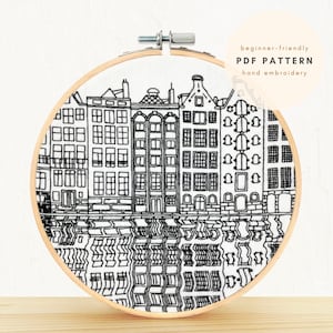 Könnte beinhalten: Schwarzweißes Hand-Stickmuster einer Stadtlandschaft mit Gebäuden, die sich im Wasser spiegeln. Das Muster ist auf einem weißen Stoff in einem hölzernen Stickrahmen. Der Text "beginner-friendly PDF PATTERN hand embroidery" befindet sich in einem Kreis oben rechts.