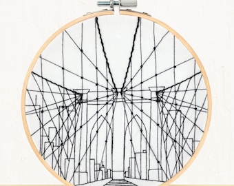 Op de afbeelding: Zwart-witte borduurringkunst die de Brooklyn Bridge met een stadsbeeld op de achtergrond afbeeldt.