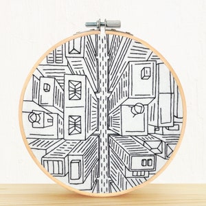 Op de afbeelding: Zwart-witte borduurringkunst die een skyline vanuit vogelperspectief toont. De gebouwen zijn met zwarte draad omrand op een witte stoffen achtergrond.