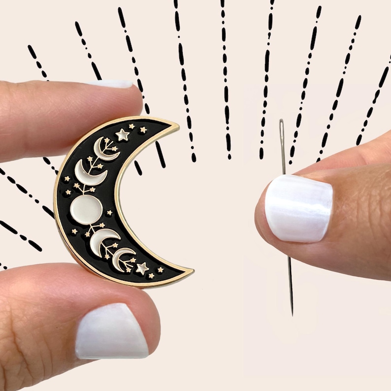 May include: Black and gold crescent moon enamel pin with phases of the moon and stars. The pin is held between two fingers. A needle is held in the other hand. The background has black dashes.