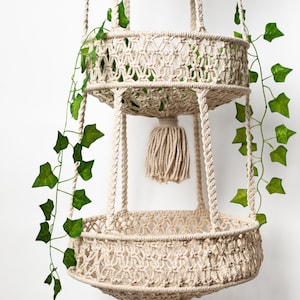 Puede incluir: Un macetero de macramé de dos niveles con una cuerda de color crema y borlas. El macetero está decorado con hiedra verde.