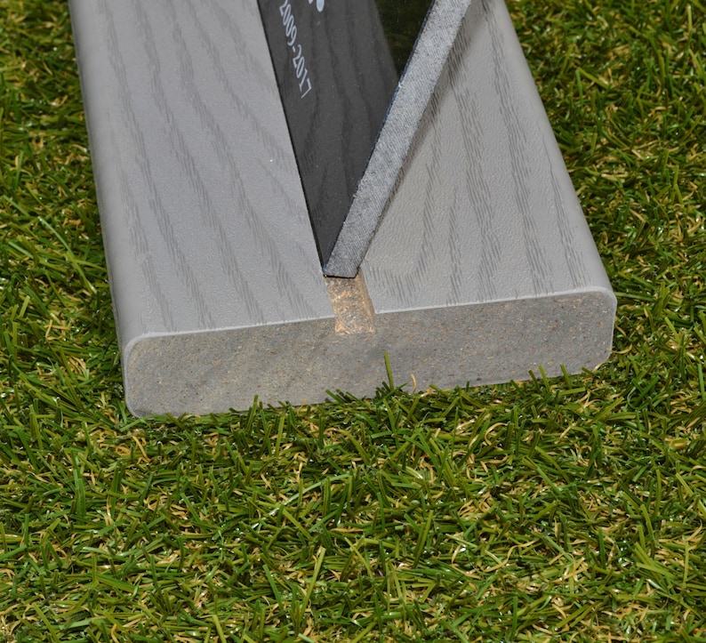 May include: Close-up of a gray wooden sign holder with a black sign inserted. The sign holder has a groove in the center and is sitting on a green artificial grass surface.