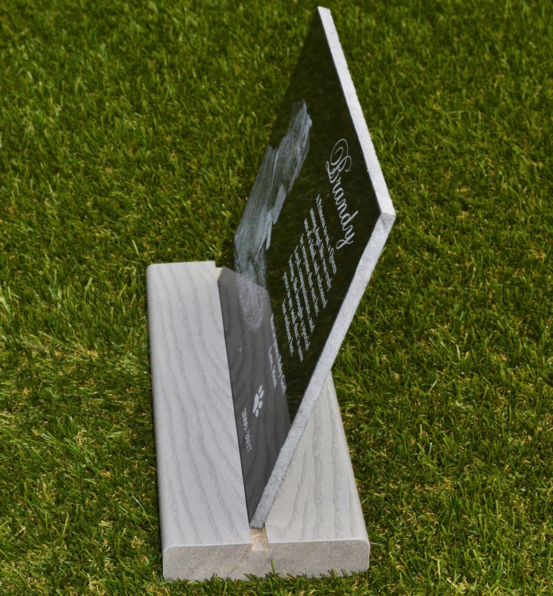 May include: Black granite pet memorial plaque with a paw print and the name "Brandy" engraved on it. The plaque is standing on a light gray wooden stand.