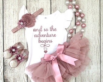 May include: A baby outfit set in blush pink with a white bodysuit that says "and so the adventure begins" in rose gold glitter. The set includes a tulle skirt, a headband with blush pink flowers, and a pair of shoes with blush pink flowers.