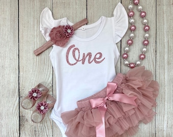May include: A baby girl's first birthday outfit with a white bodysuit that says "One" in rose gold glitter, a blush pink tulle tutu skirt with a matching satin bow, a rose gold headband with a flower, and a blush pink and white beaded necklace.