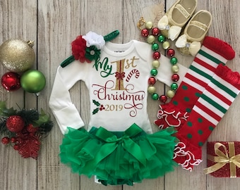 May include: A white long-sleeved onesie with the text "My 1st Christmas 2019" in gold glitter lettering. The onesie is paired with a green tulle tutu, a red and white striped leg warmer, a gold beaded necklace, and gold shoes with white bows.