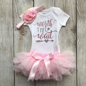 Puede incluir: Un tut&uacute; rosa con un body blanco que dice "worth the wait" en purpurina dorada rosa con una flecha y un coraz&oacute;n en purpurina plateada. El body tiene una diadema de flores rosas con un strass.