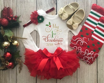 May include: A festive baby outfit for a first Christmas. The set includes a white long-sleeved onesie with "My 1st Christmas 2018" in gold and red script, a red tutu, gold shoes, a headband, and red and green striped socks.