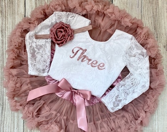May include: A white lace bodysuit with long sleeves and the word "Three" in rose gold glitter on the front. The bodysuit is paired with a dusty rose tulle skirt and a rose gold glitter headband with a rose accent.