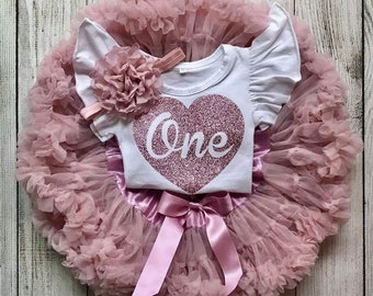 May include: A white onesie with a pink glitter heart that says "One" on it. The onesie has ruffled sleeves and is paired with a dusty rose tulle tutu with a pink satin bow.