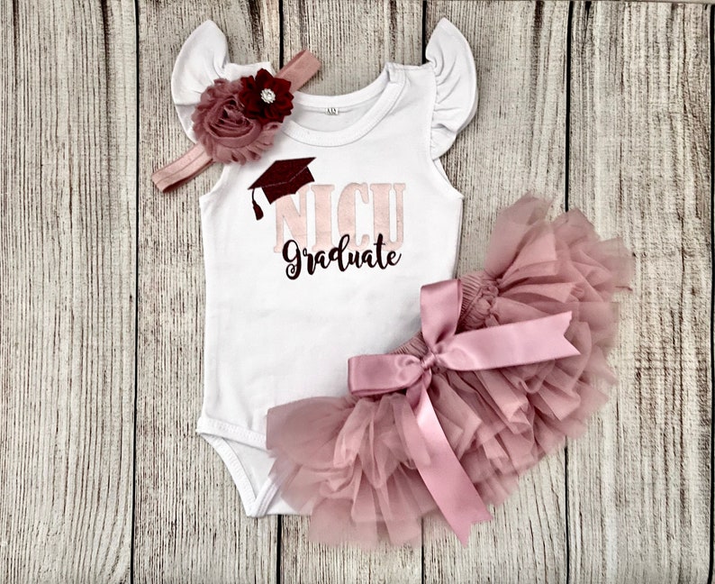May include: A white onesie with a pink and maroon graduation cap and the text "NICU Graduate". The onesie is paired with a pink tulle tutu and a pink headband with a maroon flower.