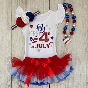 May include: A white bodysuit with red, white, and blue glitter accents. The bodysuit says "My First 4th of July" with a red, white, and blue heart. The bodysuit is paired with a red, white, and blue tulle tutu and a red, white, and blue beaded necklace.