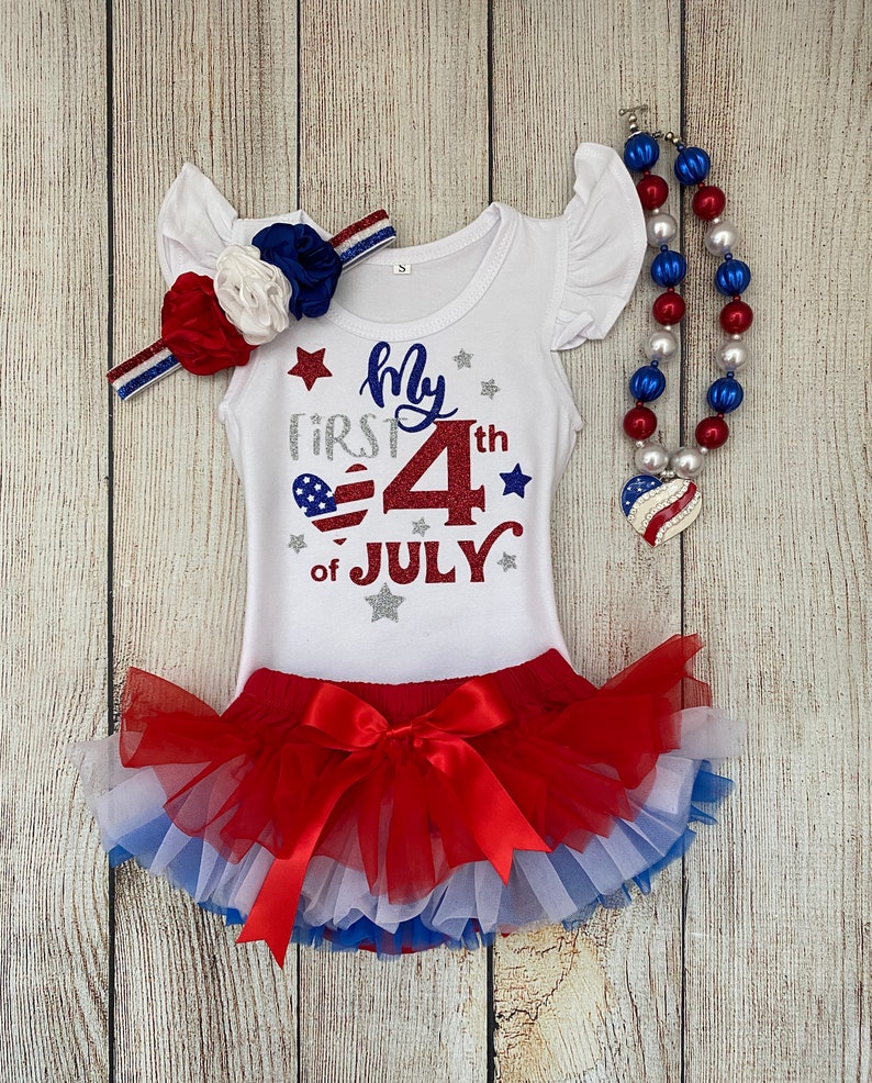 May include: A white bodysuit with red, white, and blue glitter accents. The bodysuit says "My First 4th of July" with a red, white, and blue heart. The bodysuit is paired with a red, white, and blue tulle tutu and a red, white, and blue beaded necklace.