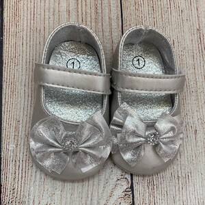May include: A pair of silver baby shoes with a bow on the front. The shoes are size 1.