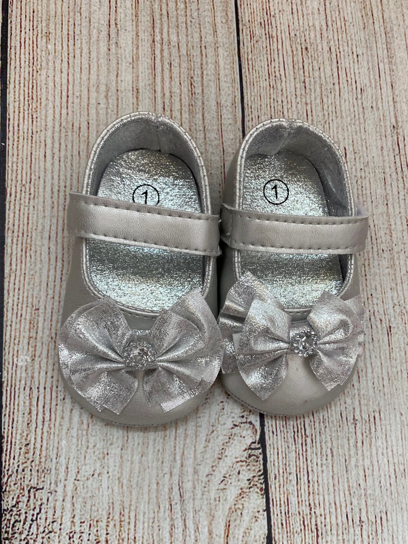 May include: A pair of silver baby shoes with a bow on the front. The shoes are size 1.