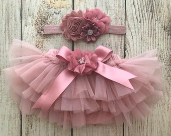 May include: A dusty rose tulle tutu with a matching satin bow and flower embellishment. A dusty rose headband with a flower embellishment is also included.