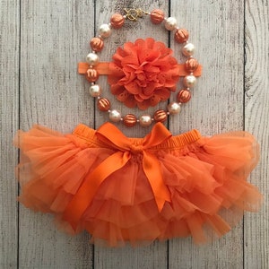 Peut inclure: Un ensemble de b&eacute;b&eacute; compos&eacute; d'une culotte en tulle orange, d'un bandeau assorti avec une fleur orange et d'un collier de perles orange et blanches.