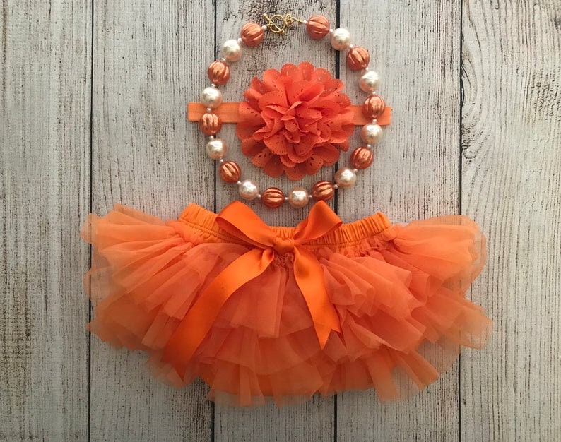 Peut inclure: Un ensemble de b&eacute;b&eacute; compos&eacute; d'une culotte en tulle orange, d'un bandeau assorti avec une fleur orange et d'un collier de perles orange et blanches.