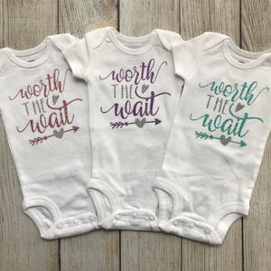 Puede incluir: Tres bodies blancos para beb&eacute; con el texto "worth the wait" en letras de purpurina. Cada body tiene un color de purpurina diferente: rosa, morado y turquesa.